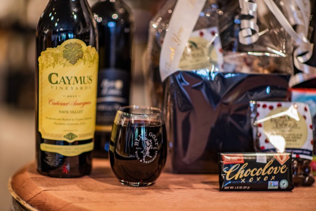 A full glass of wine, sitting in front of a Blue Goat gift basket and assorted goodies.